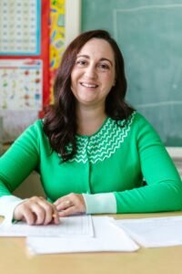 Engaging image of a smiling teacher seated at a desk in a classroom environment.